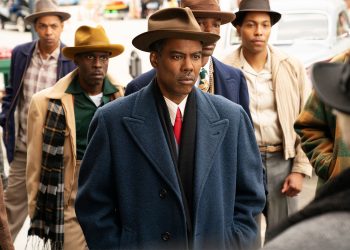 Chris Rock como o personagem Loy Cannon na quarta temporada da série Fargo, usando um casaco azul e chapéu, com outros homens ao fundo em uma cena de rua.