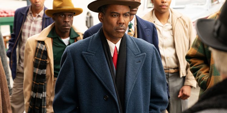 Chris Rock como o personagem Loy Cannon na quarta temporada da série Fargo, usando um casaco azul e chapéu, com outros homens ao fundo em uma cena de rua.