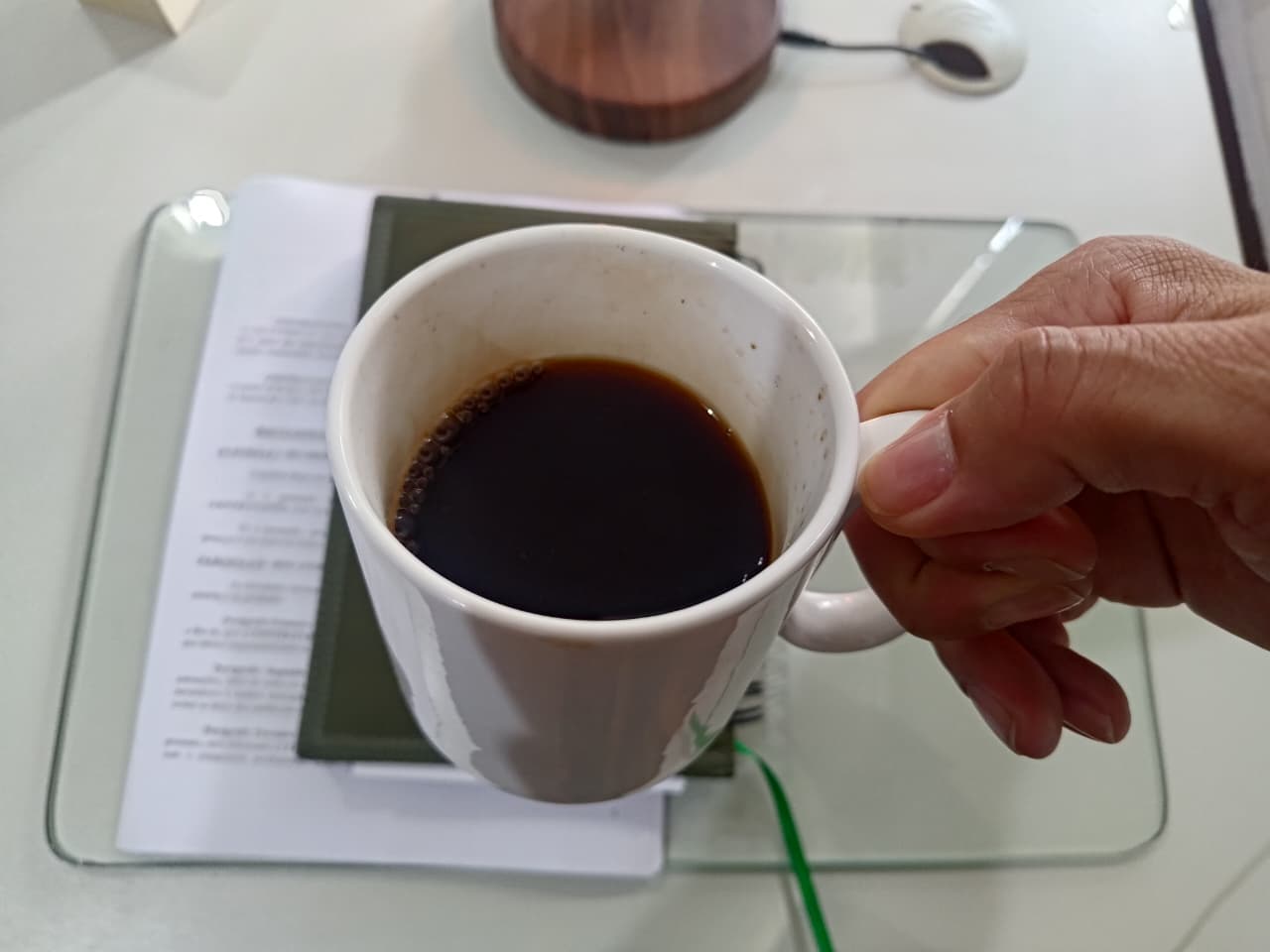 Uma mão segura uma xícara branca com café preto, sobre uma mesa com papéis e um caderno ao fundo.