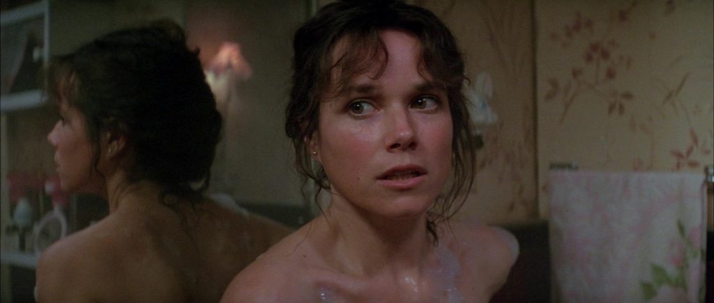 A atriz Barbara Hershey como Carla Moran no filme 'O Enigma do Mal', olhando assustada para algo fora de quadro dentro de um banheiro.