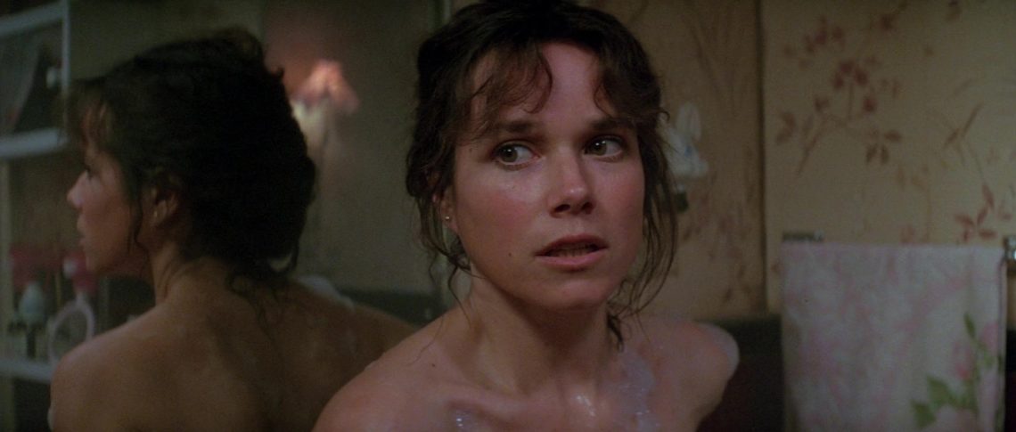 A atriz Barbara Hershey como Carla Moran no filme 'O Enigma do Mal', olhando assustada para algo fora de quadro dentro de um banheiro.