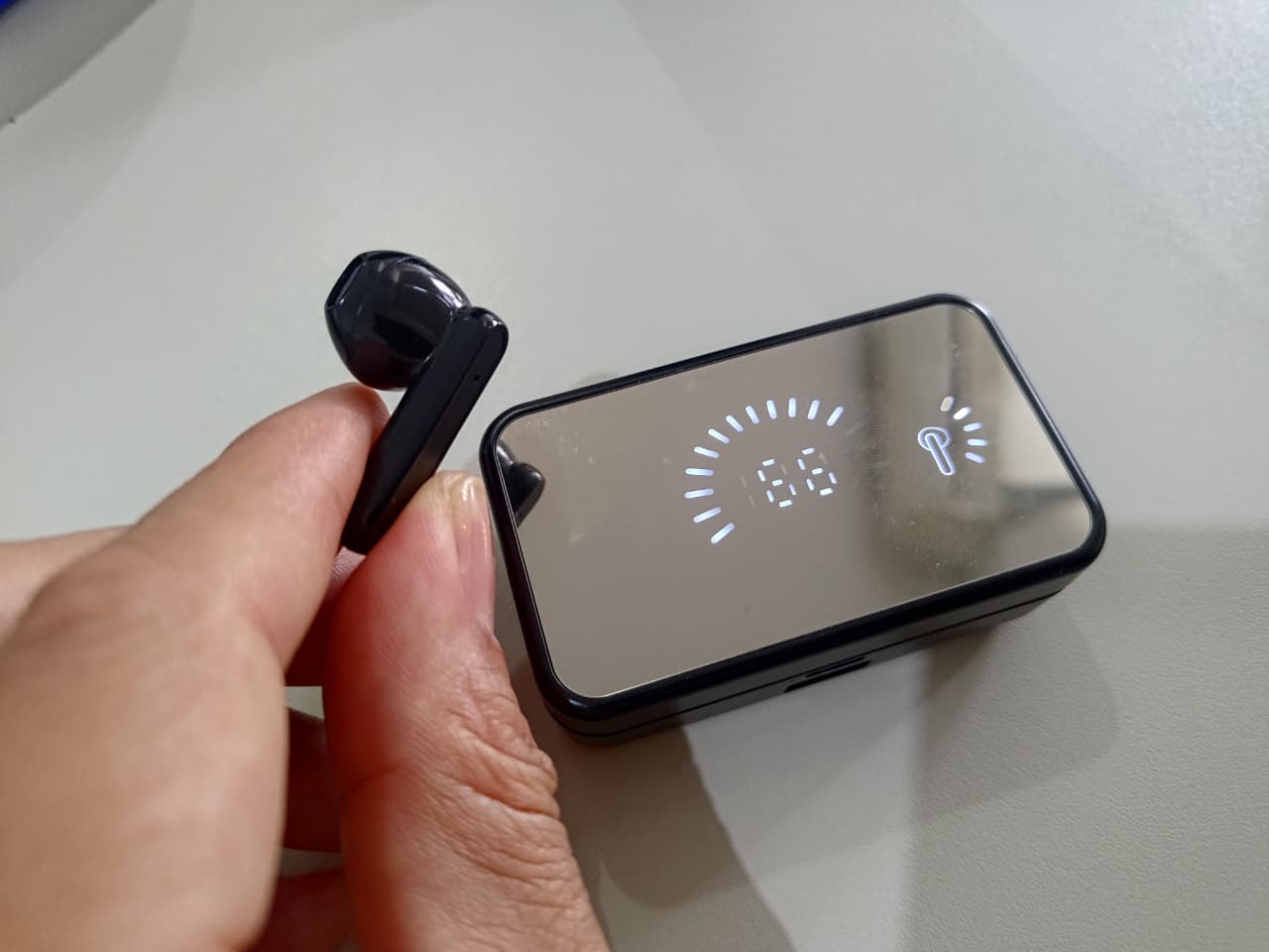 Mão segurando um fone de ouvido sem fio preto, com a caixa carregadora espelhada ao fundo mostrando o nível de bateria.