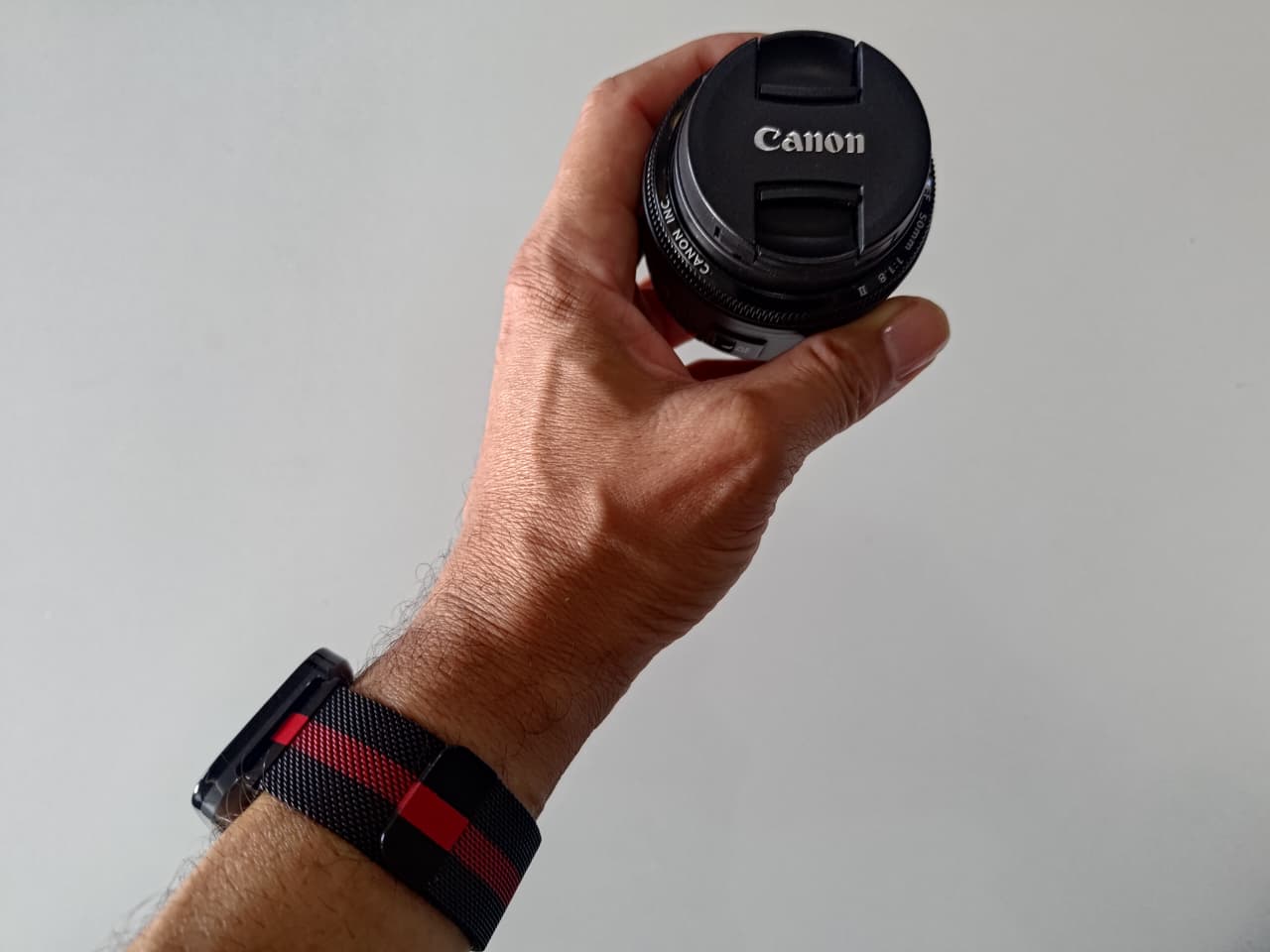Uma mão segura uma lente Canon 50mm com a tampa protetora, enquanto no pulso se vê um smartwatch com pulseira preta e vermelha.