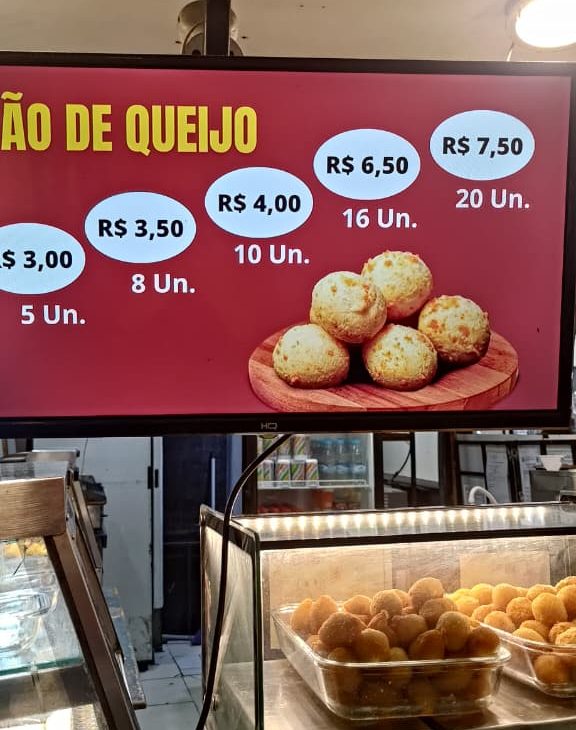 Menu digital de uma lanchonete exibindo os preços de porções de pão de queijo, com uma vitrine de salgados abaixo.