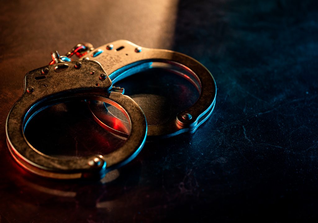 Close-up de um par de algemas de metal sobre uma superfície escura, dramaticamente iluminadas por uma luz que transita entre o vermelho e o azul.