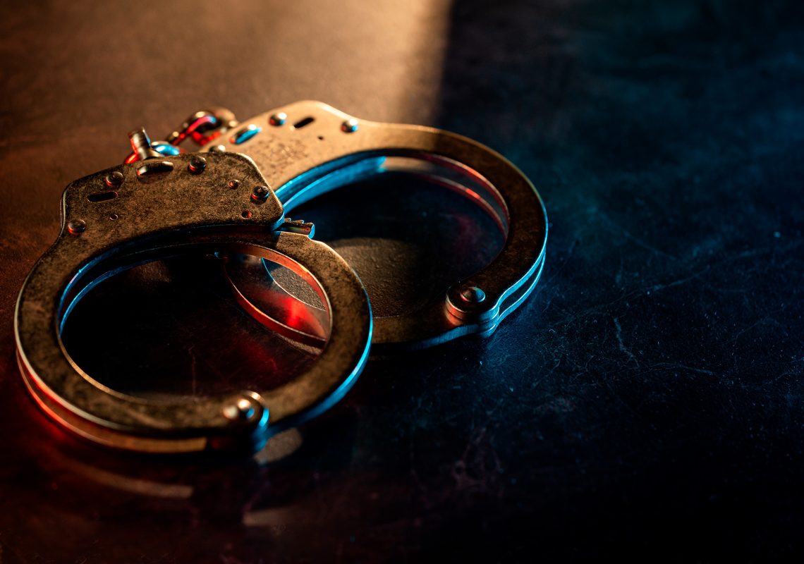 Close-up de um par de algemas de metal sobre uma superfície escura, dramaticamente iluminadas por uma luz que transita entre o vermelho e o azul.