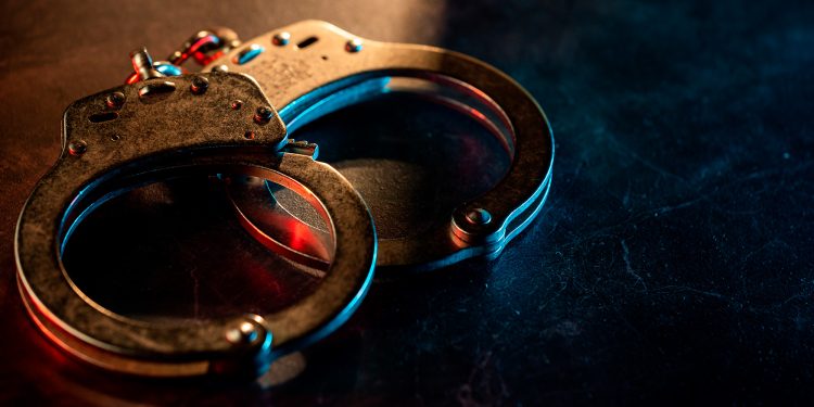 Close-up de um par de algemas de metal sobre uma superfície escura, dramaticamente iluminadas por uma luz que transita entre o vermelho e o azul.