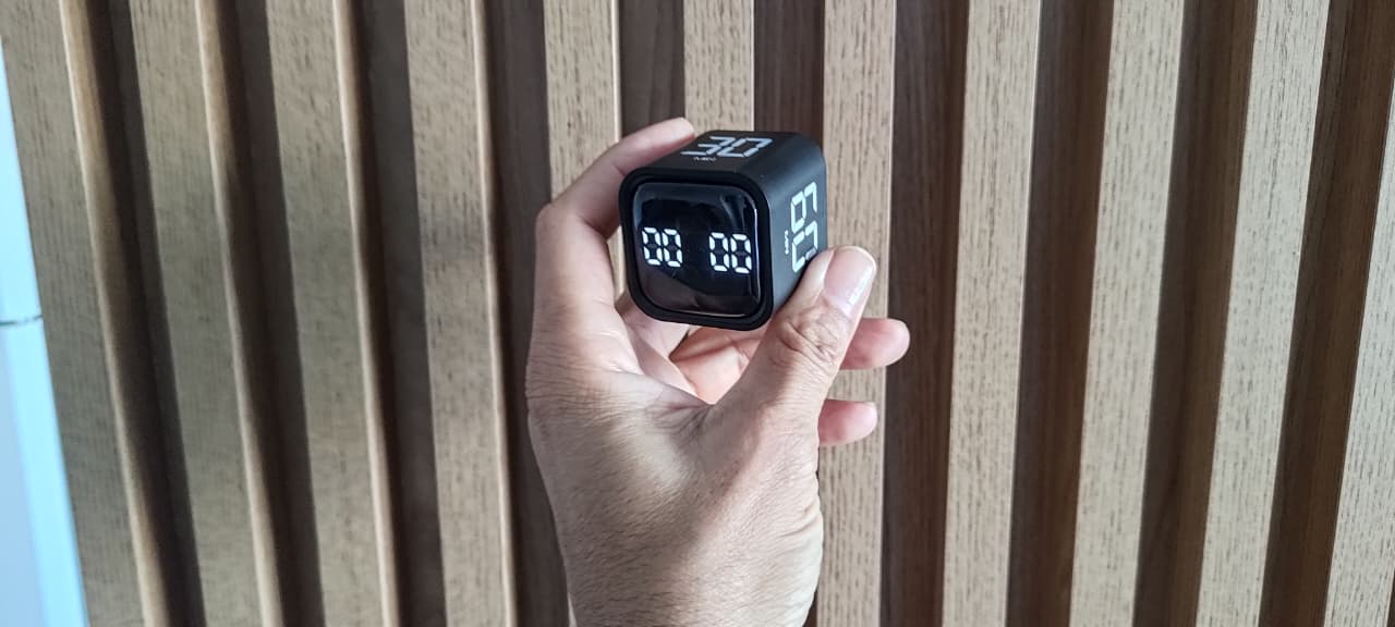 Mão segurando um timer digital preto em formato de cubo, com o display zerado, contra um fundo de painel de madeira.