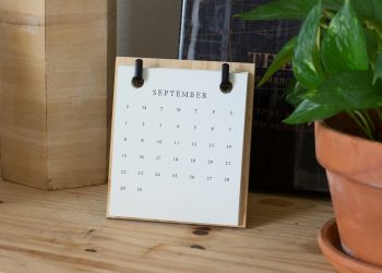 Calendário de mesa de madeira exibindo a página do mês de setembro, apoiado em uma prateleira ao lado de um vaso de planta.