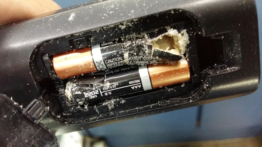 Compartimento de pilhas de um controle remoto ou aparelho eletrônico mostrando duas pilhas estouradas e vazando, com resíduos esbranquiçados.