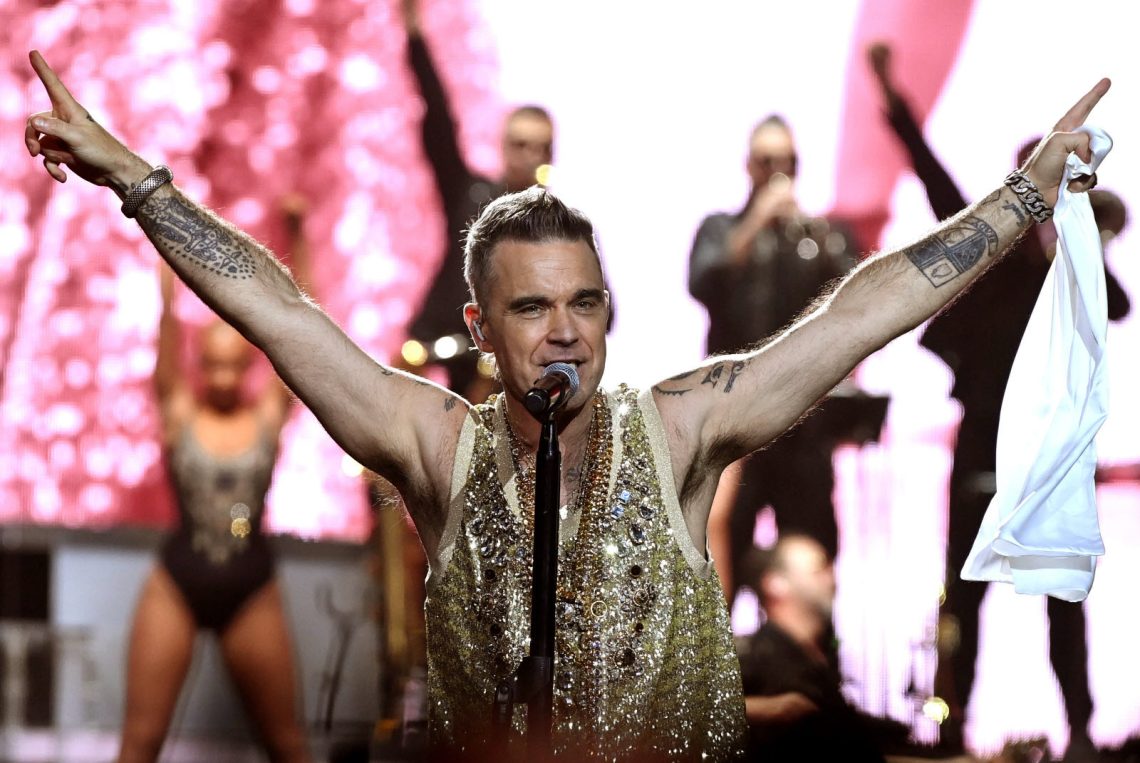 Robbie Williams no palco com os braços erguidos, vestindo um colete dourado e sorrindo para a plateia.