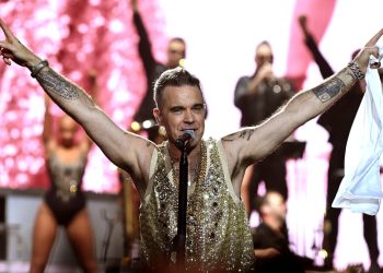 Robbie Williams no palco com os braços erguidos, vestindo um colete dourado e sorrindo para a plateia.