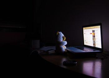 Urso de pelúcia sentado em frente a um laptop com a tela acesa em um quarto escuro à noite.
