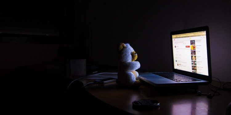 Urso de pelúcia sentado em frente a um laptop com a tela acesa em um quarto escuro à noite.