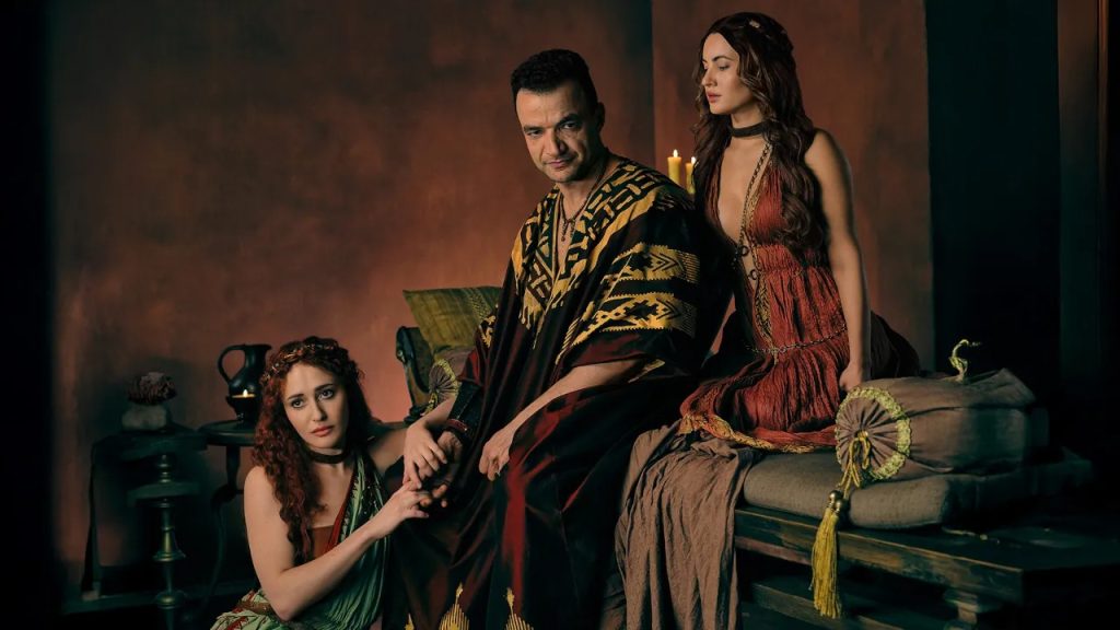 Nick E. Tarabay como Ashur sentado em um trono, ladeado por duas mulheres com vestes romanas, em uma cena promocional da série Spartacus: House of Ashur.
