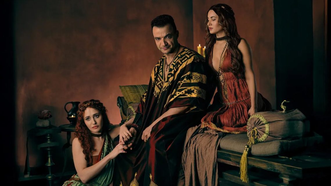 Nick E. Tarabay como Ashur sentado em um trono, ladeado por duas mulheres com vestes romanas, em uma cena promocional da série Spartacus: House of Ashur.