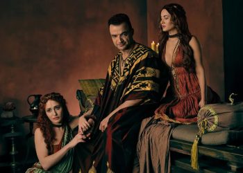 Nick E. Tarabay como Ashur sentado em um trono, ladeado por duas mulheres com vestes romanas, em uma cena promocional da série Spartacus: House of Ashur.