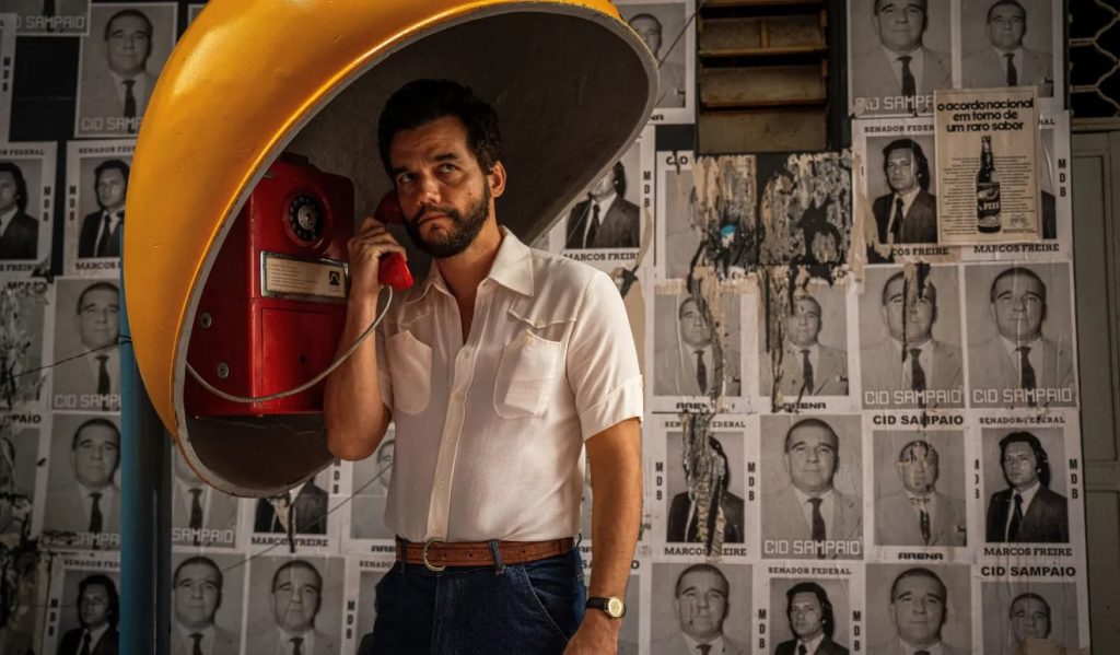 Wagner Moura caracterizado como Marcelo no filme O Agente Secreto, usando um orelhão amarelo em frente a um muro coberto de cartazes políticos da década de 1970.