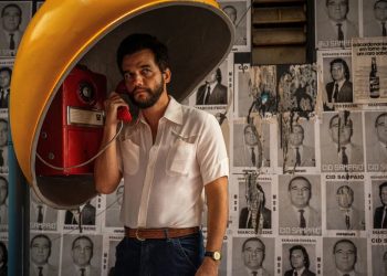 Wagner Moura caracterizado como Marcelo no filme O Agente Secreto, usando um orelhão amarelo em frente a um muro coberto de cartazes políticos da década de 1970.