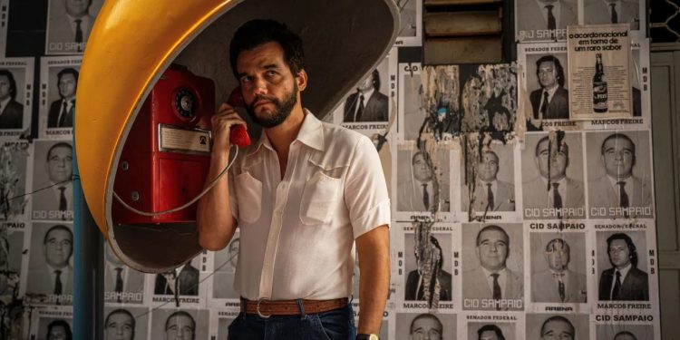 Wagner Moura caracterizado como Marcelo no filme O Agente Secreto, usando um orelhão amarelo em frente a um muro coberto de cartazes políticos da década de 1970.