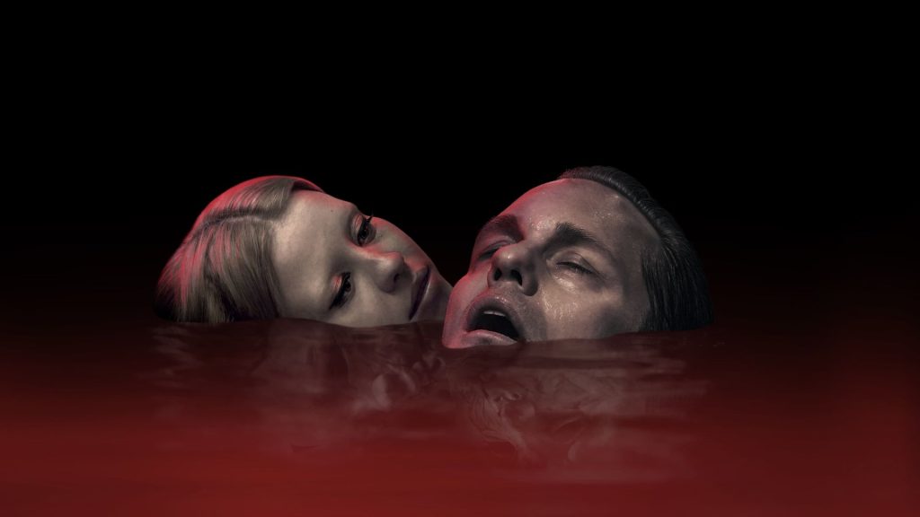 Close-up dos atores Mia Goth e Alexander Skarsgård com os rostos emergindo de um líquido vermelho escuro, em uma cena do filme Piscina Infinita. Título da Imagem: Mia Goth e Alexander Skarsgård - Piscina Infinita