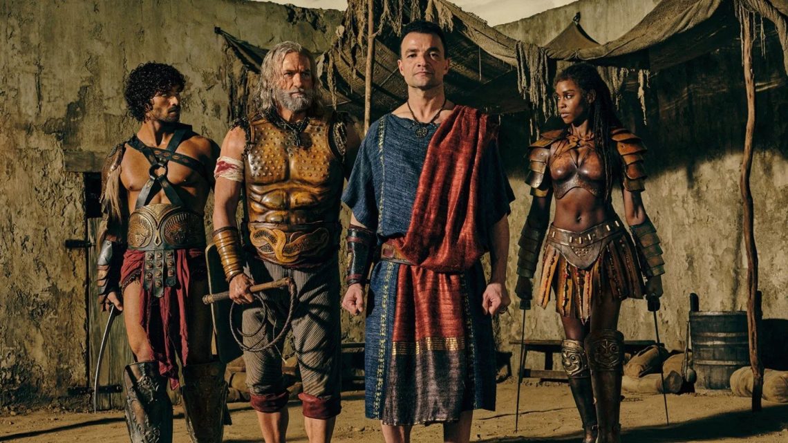 Personagens de Spartacus House of Ashur em trajes de batalha e togas romanas em um cenário de arena rústico.