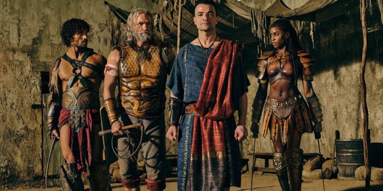 Personagens de Spartacus House of Ashur em trajes de batalha e togas romanas em um cenário de arena rústico.