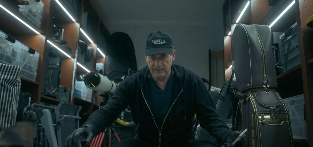 Uma cena fotorrealista em close-up de um homem (Jon Hamm) agachado em uma garagem de luxo bem iluminada por fitas de LED, cercado por prateleiras com equipamentos caros: um telescópio astronômico profissional, bolsas de golfe HONMA com detalhes dourados, racks de equipamentos e caixas de plástico. O homem veste um moletom preto, luvas de couro e um boné "Larsen's Menemsha", e segura uma ferramenta metálica. Uma ilustração perfeita do acúmulo de bens caros mencionado no artigo.