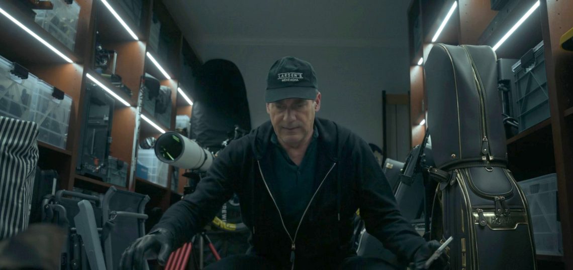 Uma cena fotorrealista em close-up de um homem (Jon Hamm) agachado em uma garagem de luxo bem iluminada por fitas de LED, cercado por prateleiras com equipamentos caros: um telescópio astronômico profissional, bolsas de golfe HONMA com detalhes dourados, racks de equipamentos e caixas de plástico. O homem veste um moletom preto, luvas de couro e um boné "Larsen's Menemsha", e segura uma ferramenta metálica. Uma ilustração perfeita do acúmulo de bens caros mencionado no artigo.