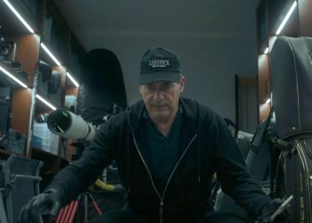 Uma cena fotorrealista em close-up de um homem (Jon Hamm) agachado em uma garagem de luxo bem iluminada por fitas de LED, cercado por prateleiras com equipamentos caros: um telescópio astronômico profissional, bolsas de golfe HONMA com detalhes dourados, racks de equipamentos e caixas de plástico. O homem veste um moletom preto, luvas de couro e um boné "Larsen's Menemsha", e segura uma ferramenta metálica. Uma ilustração perfeita do acúmulo de bens caros mencionado no artigo.
