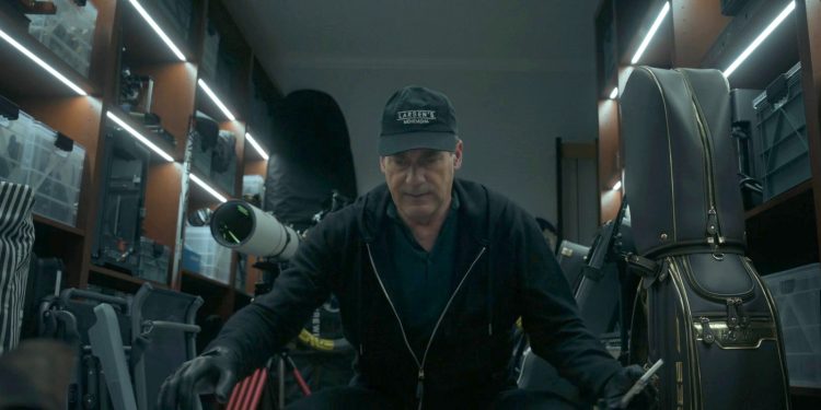 Uma cena fotorrealista em close-up de um homem (Jon Hamm) agachado em uma garagem de luxo bem iluminada por fitas de LED, cercado por prateleiras com equipamentos caros: um telescópio astronômico profissional, bolsas de golfe HONMA com detalhes dourados, racks de equipamentos e caixas de plástico. O homem veste um moletom preto, luvas de couro e um boné "Larsen's Menemsha", e segura uma ferramenta metálica. Uma ilustração perfeita do acúmulo de bens caros mencionado no artigo.