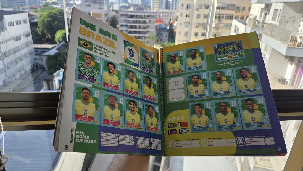 Álbum da Copa do Mundo aberto na página da Seleção Brasileira com várias figurinhas coladas, segurado por uma pessoa em frente a uma janela iluminada com vista para prédios da cidade.
