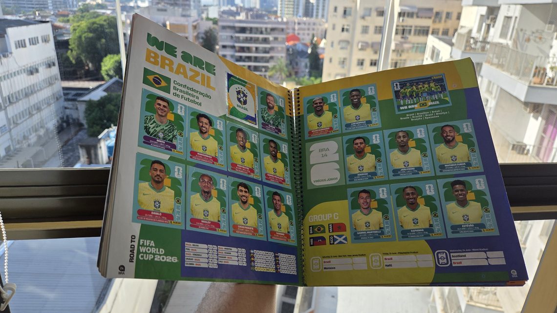 Álbum da Copa do Mundo aberto na página da Seleção Brasileira com várias figurinhas coladas, segurado por uma pessoa em frente a uma janela iluminada com vista para prédios da cidade.