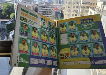 Álbum da Copa do Mundo aberto na página da Seleção Brasileira com várias figurinhas coladas, segurado por uma pessoa em frente a uma janela iluminada com vista para prédios da cidade.