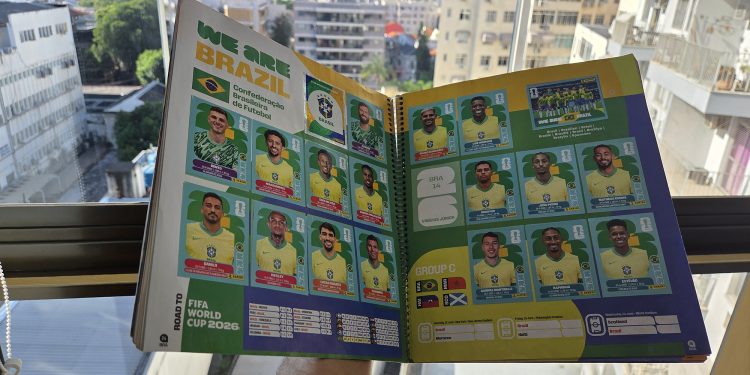 Álbum da Copa do Mundo aberto na página da Seleção Brasileira com várias figurinhas coladas, segurado por uma pessoa em frente a uma janela iluminada com vista para prédios da cidade.