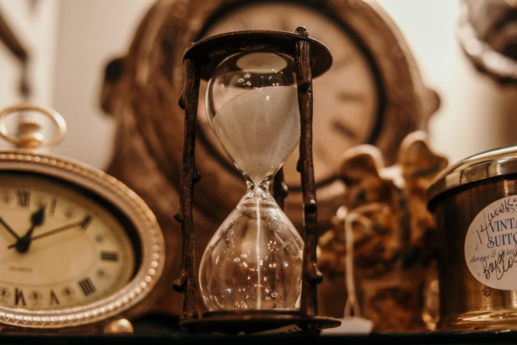 Close-up fotorrealista de uma ampulheta vintage com areia branca escorrendo, cercada por relógios antigos e objetos de época desfocados em uma loja de antiguidades.
