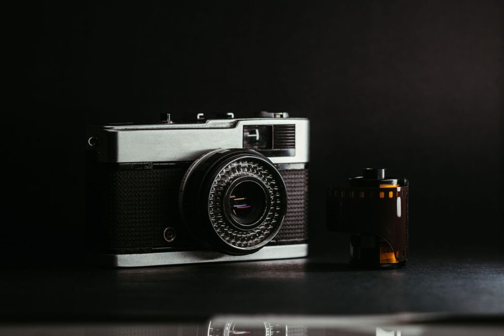 Fotografia em close de uma câmera fotográfica analógica vintage prateada e preta ao lado de um rolo de filme 35mm, com fundo escuro e iluminação dramática.