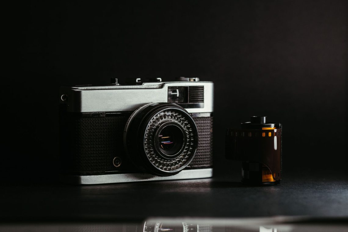 Fotografia em close de uma câmera fotográfica analógica vintage prateada e preta ao lado de um rolo de filme 35mm, com fundo escuro e iluminação dramática.