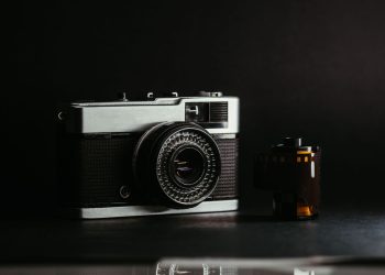Fotografia em close de uma câmera fotográfica analógica vintage prateada e preta ao lado de um rolo de filme 35mm, com fundo escuro e iluminação dramática.