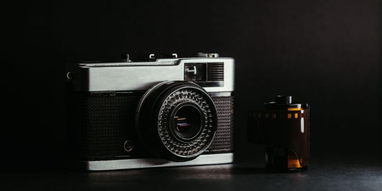 Fotografia em close de uma câmera fotográfica analógica vintage prateada e preta ao lado de um rolo de filme 35mm, com fundo escuro e iluminação dramática.