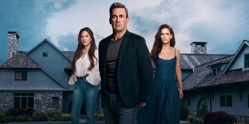 Jon Hamm de roupas escuras ao centro, acompanhado por duas mulheres, todos com expressões sérias, em pé em frente a uma grande e luxuosa mansão de subúrbio sob um céu nublado. Imagem promocional da série Seus Amigos e Vizinhos.