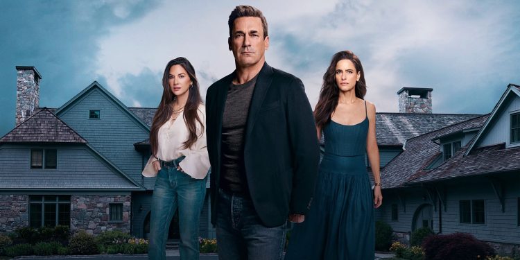 Jon Hamm de roupas escuras ao centro, acompanhado por duas mulheres, todos com expressões sérias, em pé em frente a uma grande e luxuosa mansão de subúrbio sob um céu nublado. Imagem promocional da série Seus Amigos e Vizinhos.