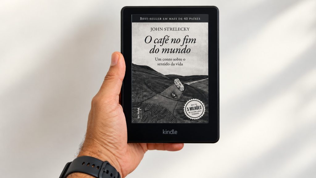 Uma mão segurando um e-reader Kindle exibindo a capa do livro O Café no Fim do Mundo, de John Strelecky.