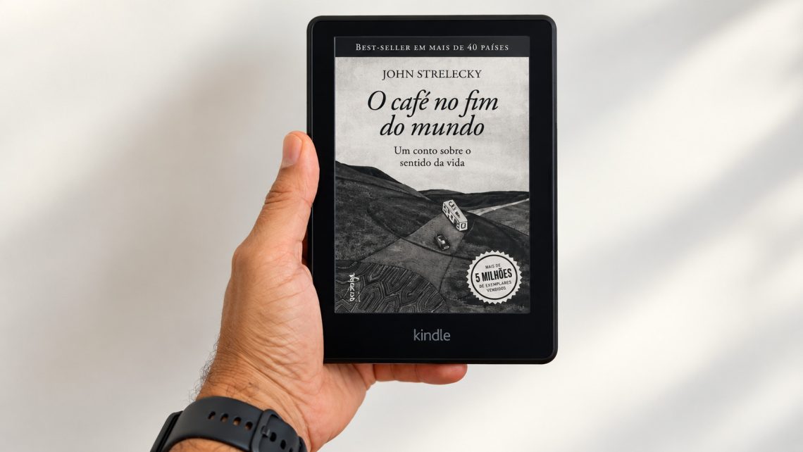 Uma mão segurando um e-reader Kindle exibindo a capa do livro O Café no Fim do Mundo, de John Strelecky.