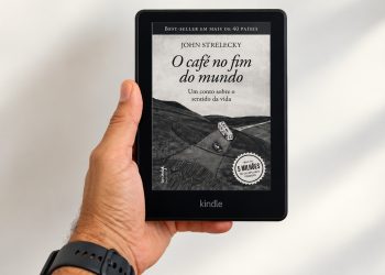 Uma mão segurando um e-reader Kindle exibindo a capa do livro O Café no Fim do Mundo, de John Strelecky.