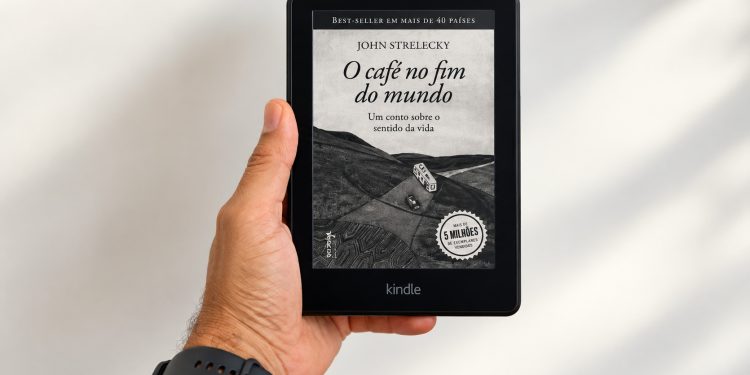 Uma mão segurando um e-reader Kindle exibindo a capa do livro O Café no Fim do Mundo, de John Strelecky.