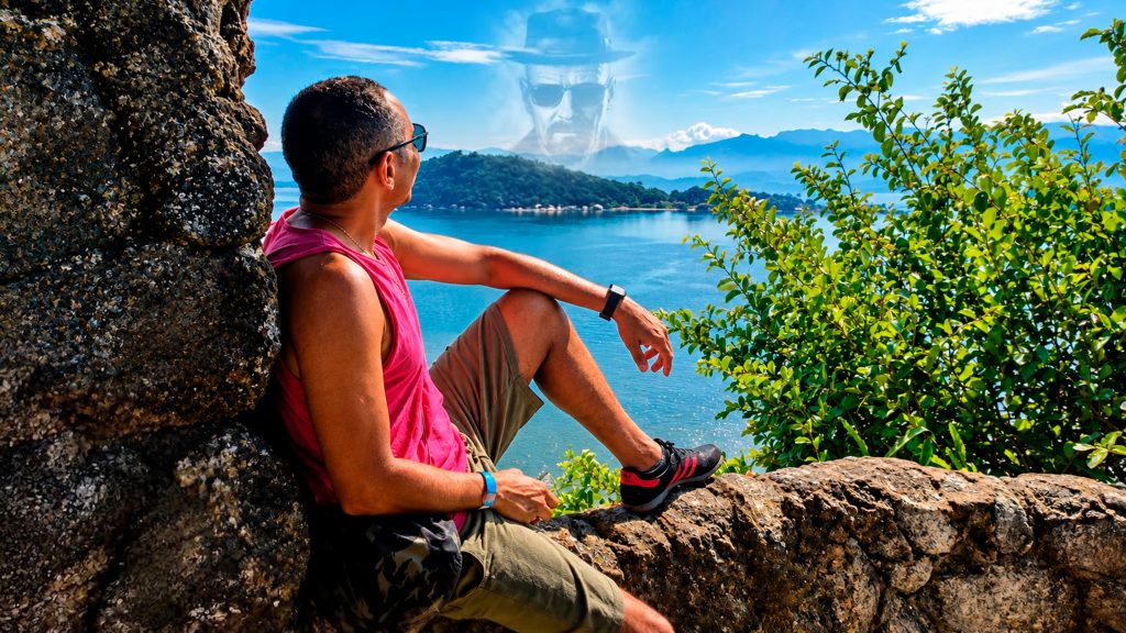 Homem sentado em um mirante de pedras observando o mar e as montanhas, com o rosto do personagem Walter White projetado sutilmente no céu azul, simbolizando a ilusão de ser o vilão.