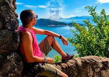 Homem sentado em um mirante de pedras observando o mar e as montanhas, com o rosto do personagem Walter White projetado sutilmente no céu azul, simbolizando a ilusão de ser o vilão.