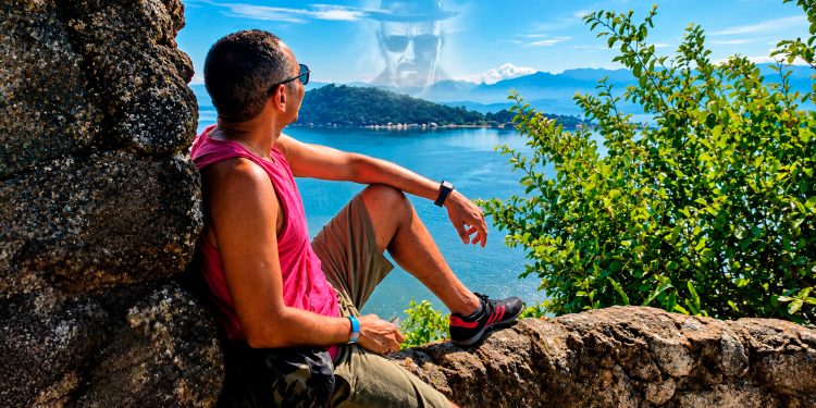 Homem sentado em um mirante de pedras observando o mar e as montanhas, com o rosto do personagem Walter White projetado sutilmente no céu azul, simbolizando a ilusão de ser o vilão.